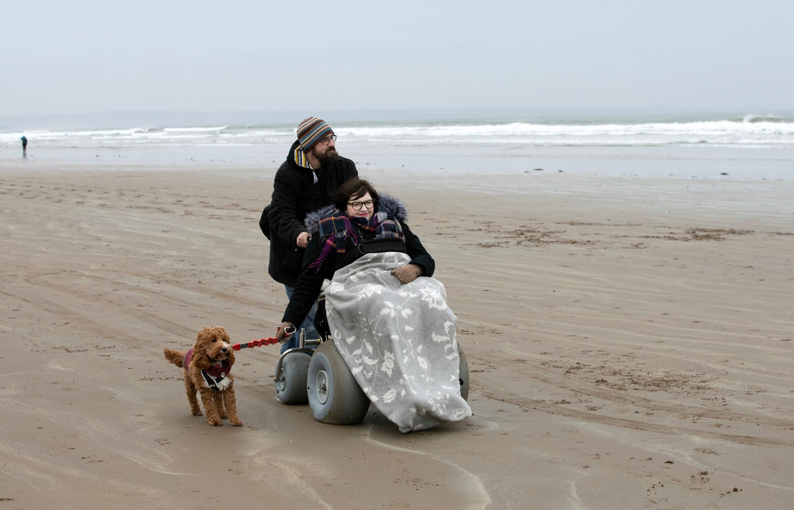 two people on the beach, with a bog. one person behind the wheel chair, and one person in one of the St Andrews Beach wheelchairs, holding the dog lead
